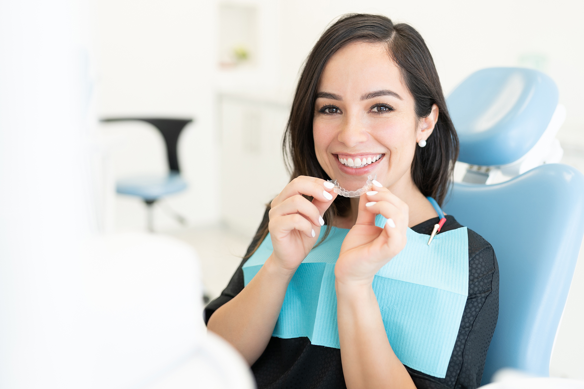 The image shows a woman with a bright smile, holding dental tools while standing in front of a dental chair, which suggests she may be a dental professional preparing for a procedure or appointment.